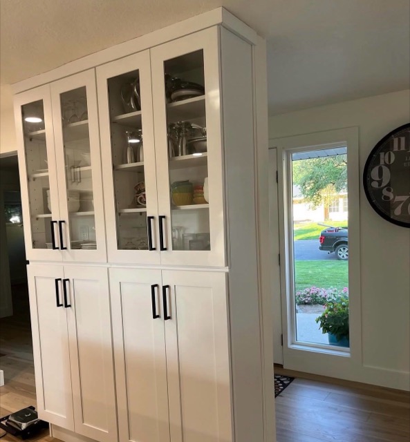 Floor-to-ceiling white shaker hutch with glass upper doors and matte black bar pulls beside the entry